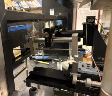 Black clamp block on metal rods mounted above a traverse stage inside the wind tunnel frame.
