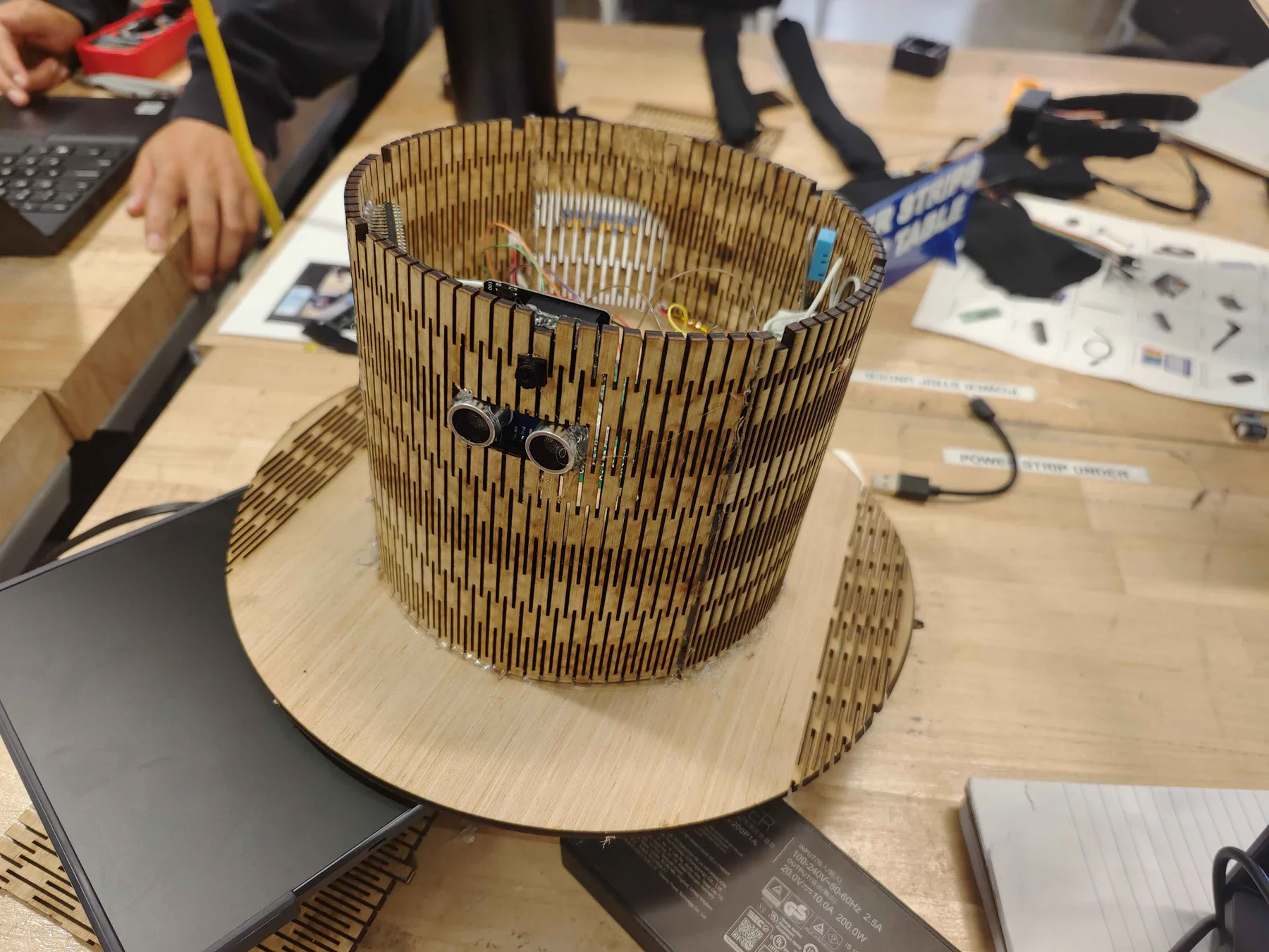 Laser-cut flexible plywood top hat with an ultrasonic sensor on the front, on a workbench.