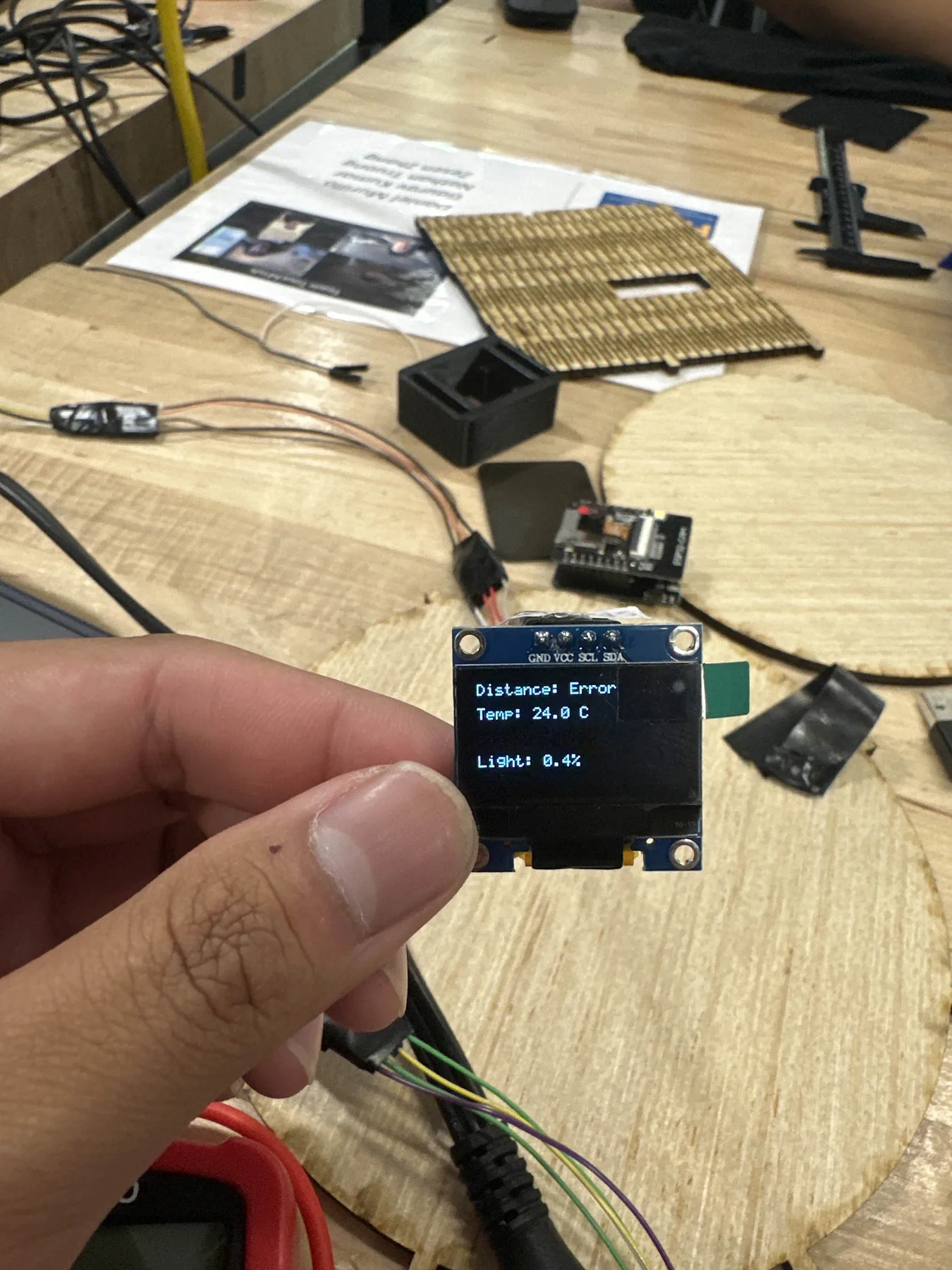 Small OLED screen held between fingers showing distance, temperature and light readings.