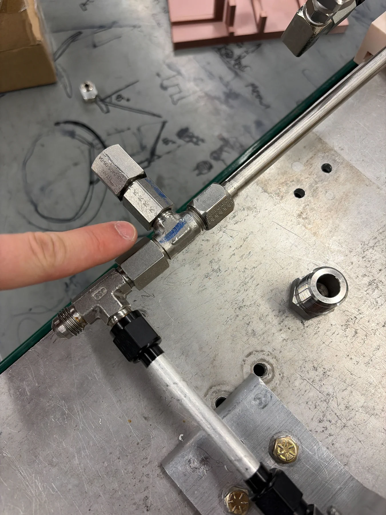 Close-up of a stainless compression tee fitting on an aluminum plate, with a finger pointing at it.