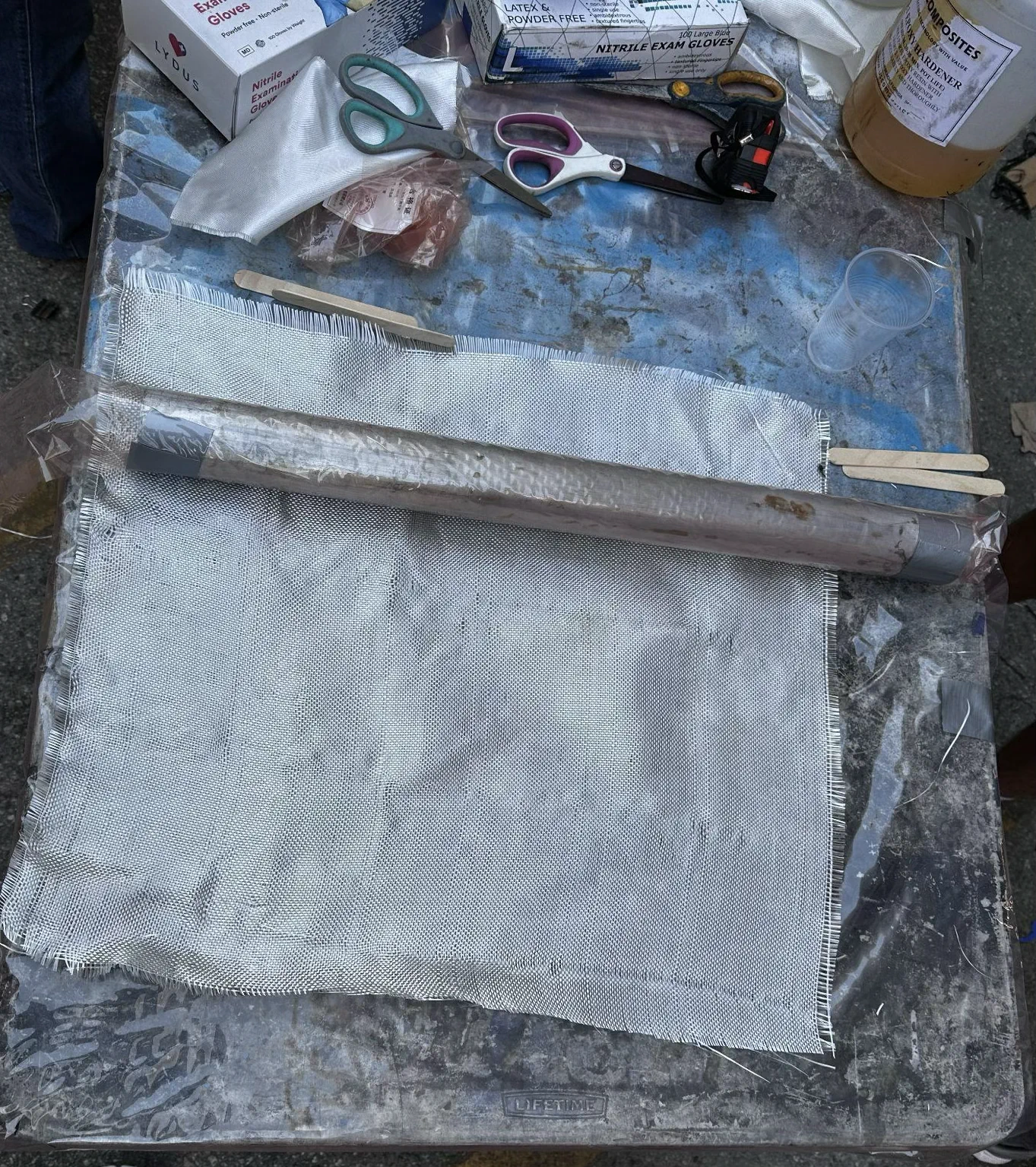 Fiberglass cloth and a wrapped steel pipe mandrel on a work table with layup tools.