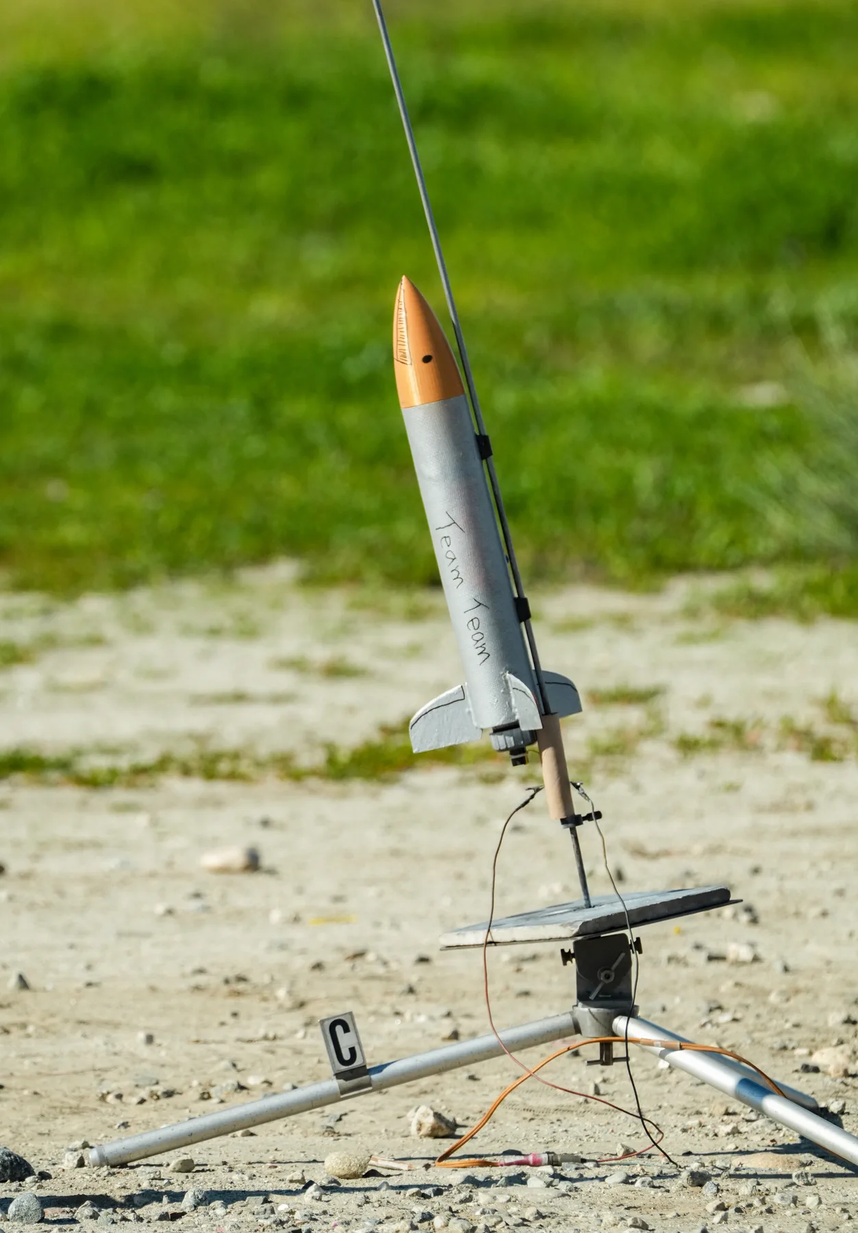 Silver model rocket labeled Team Team on a launch rail in a dirt field.