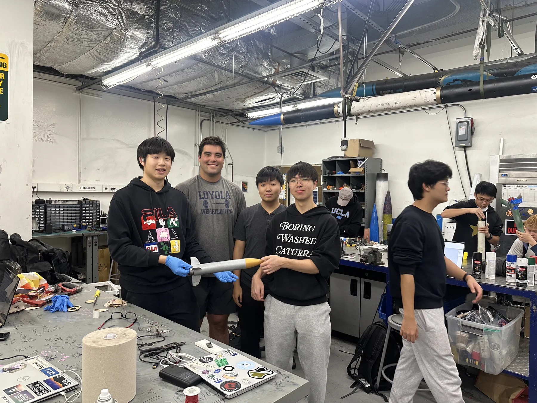 Four students in a lab holding a silver model rocket with a gold nose cone.